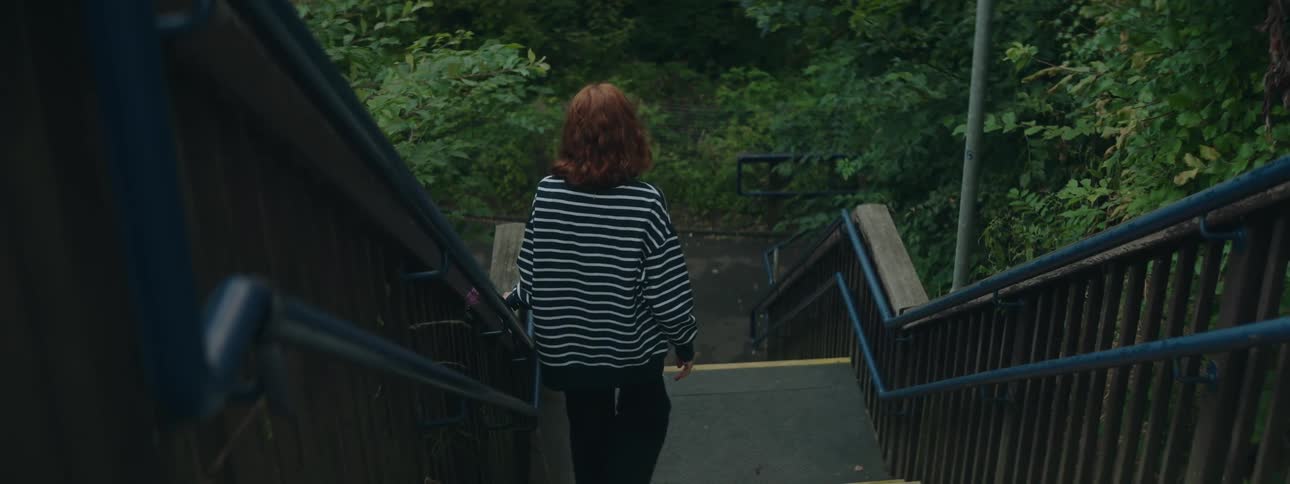 A young woman with red hair walks down a staircase leading to a lush green park