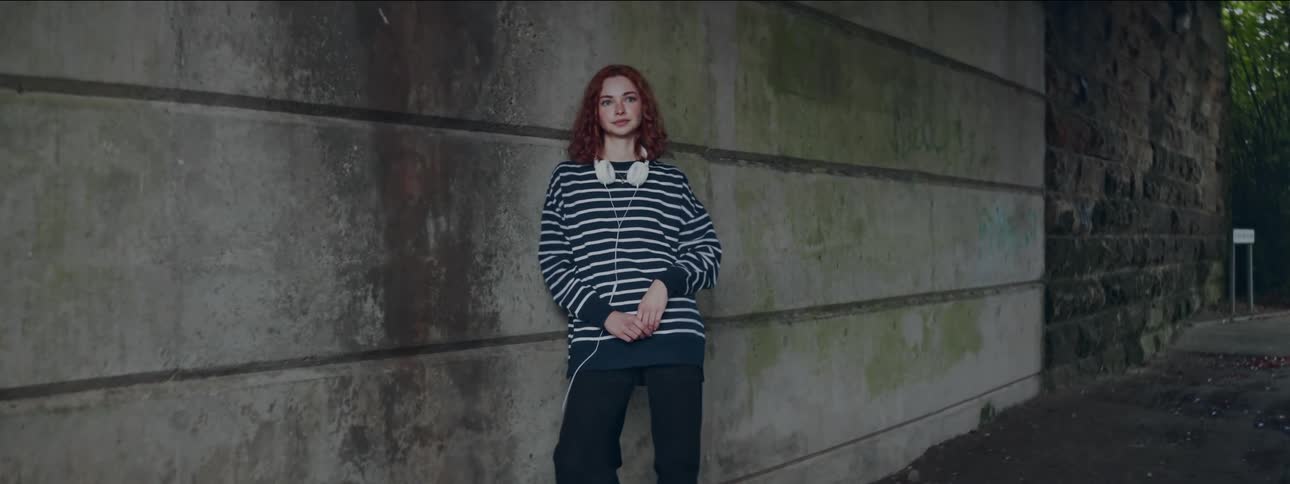 A young woman in a striped sweater leans against a concrete wall