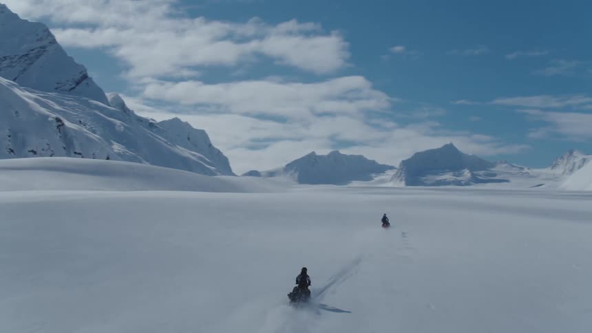 Three snowmobiles driving on a vast snowy landscape with mountains in the distance
