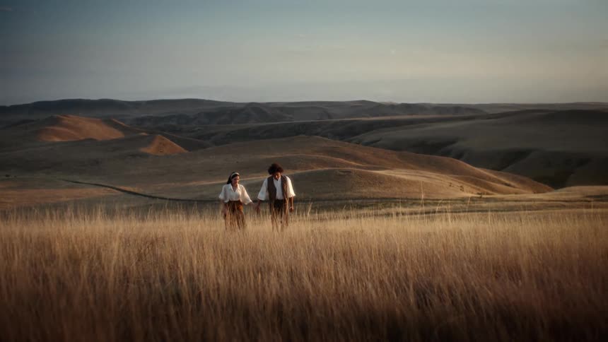Man and woman holding hands walking together through a field with hills in the background