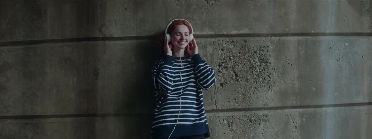 A red haired girl leans on a wall and smiles while listening to headphones