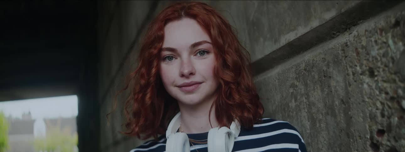 A young woman with red hair and white headphones around her neck smiles while leaning against an underpass wall