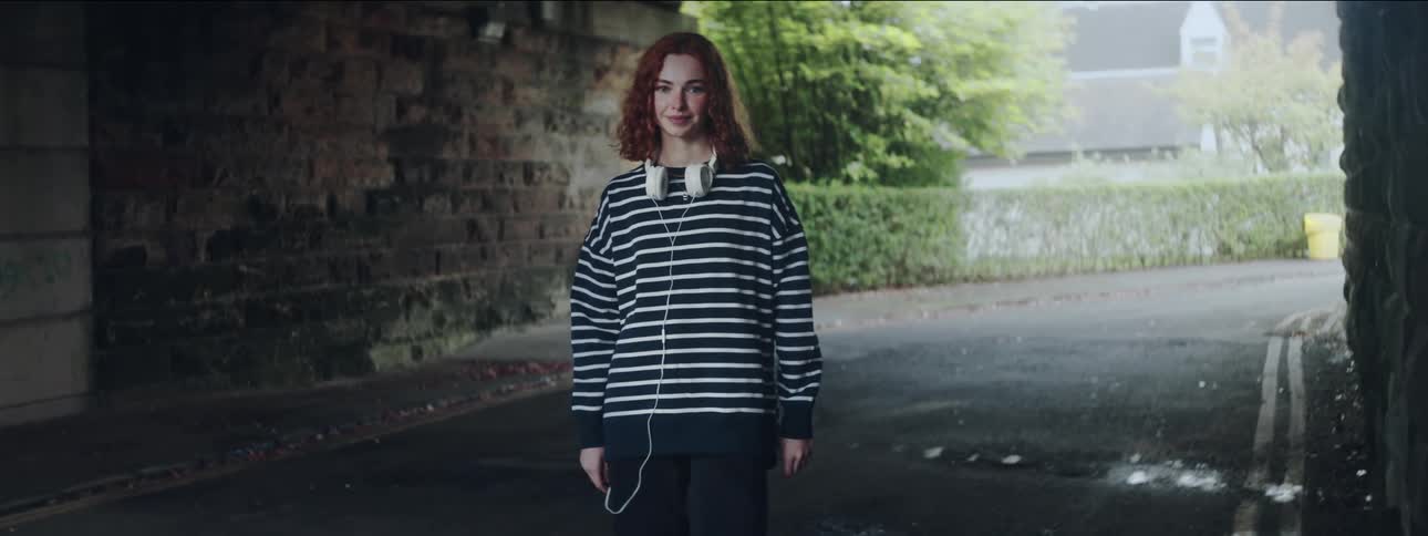 A woman with red curly hair wearing a striped shirt and white headphones stands on a road under an overpass