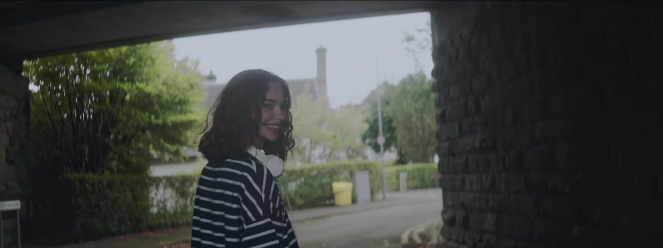 A young woman with red hair walking out of an underpass then turning her head back with a smile