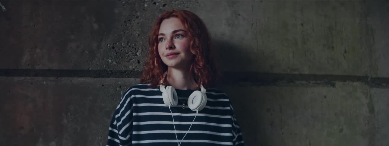 Girl with red hair wearing striped shirt leaning on wall looking off to the side