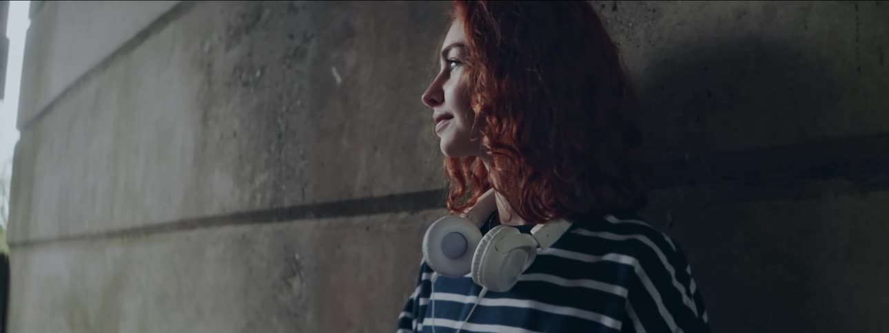Girl with red hair wearing striped shirt leaning against cement wall outdoors