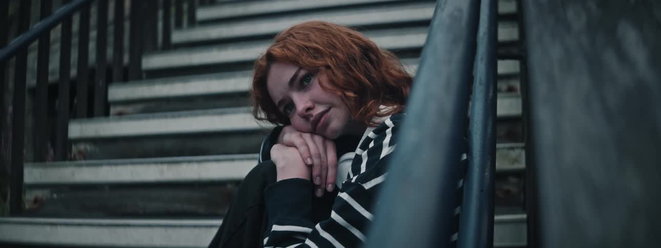 A redheaded young woman sitting in a staircase with a sad smile resting her head on her hands and her hands on her knee