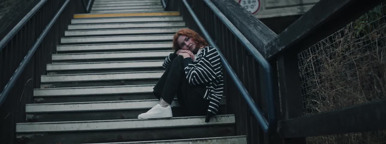 A red haired girl leans her head against her hands resting on her knees while sitting on a staircase