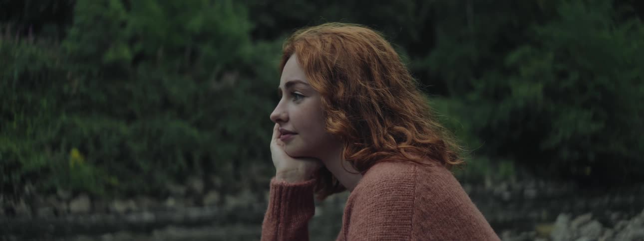A redheaded young woman in a park by a river smiling while resting her face on her hand