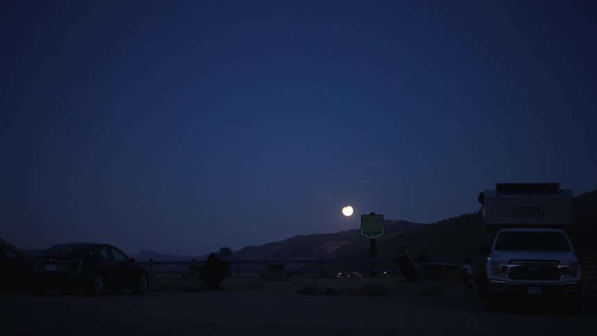 The moon hangs over a mountain range near a lot with vehicles parked in it