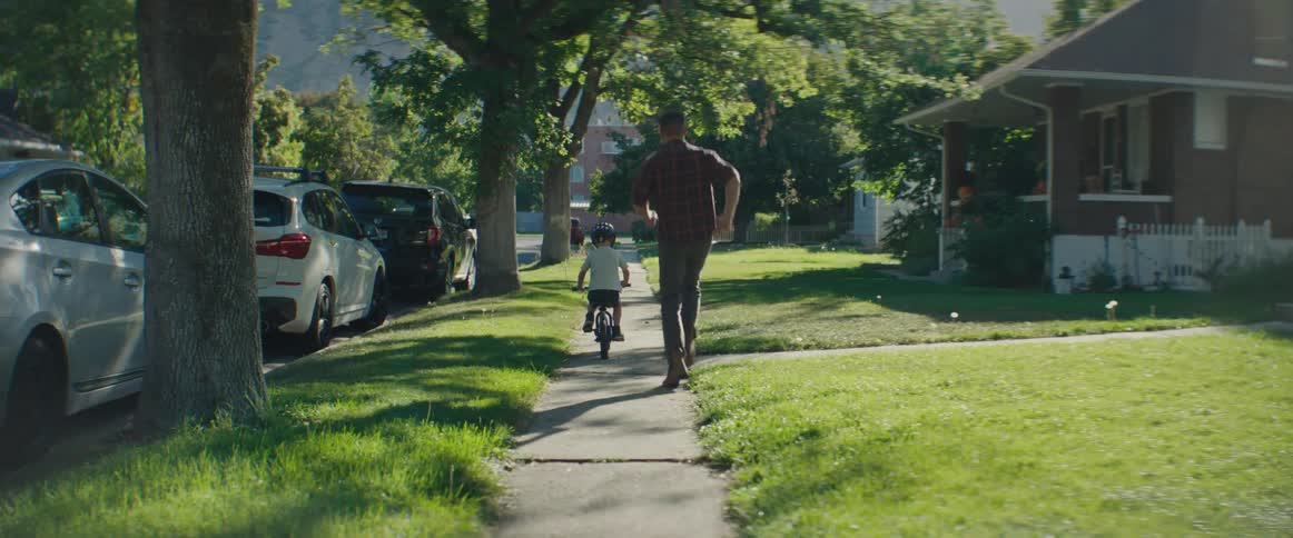 A father runs alongside his son who is riding a bike down a suburban street