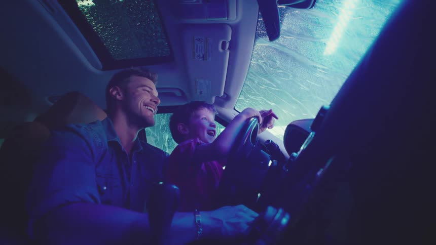 The son is sitting on his fathers lap while they drive in the rain