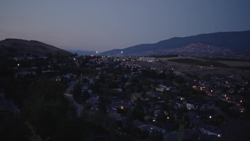 A city nestled in a valley between mountains with lights twinkling from houses and streetlights illuminating the roads under the twilight sky