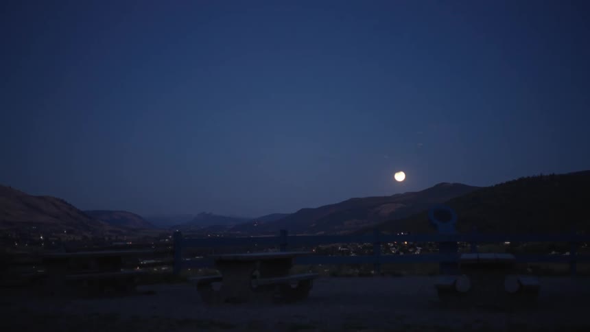 A place with three empty benches and a view of a small town at night with moonlight in the sky