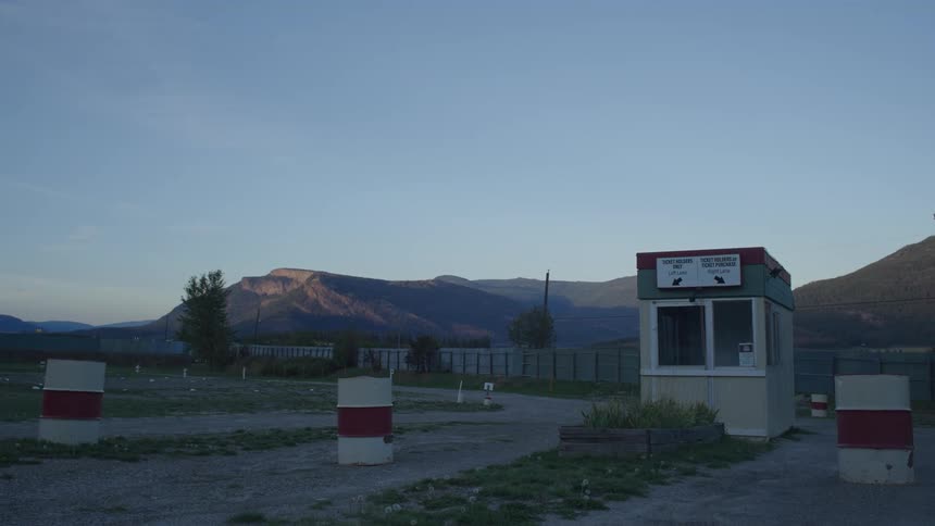 Barrels blocking the entrance to a ticket booth at an abandoned drive in theater with mountains behind