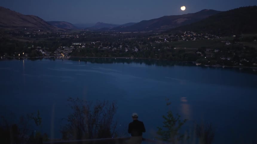 Man stands across the water from a town sitting at the base of the mountains under a full moon at night