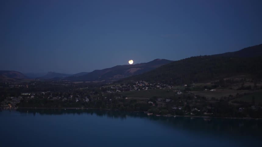 Mountain landscape with scattered houses on the edge of a lake and the moon on the horizon