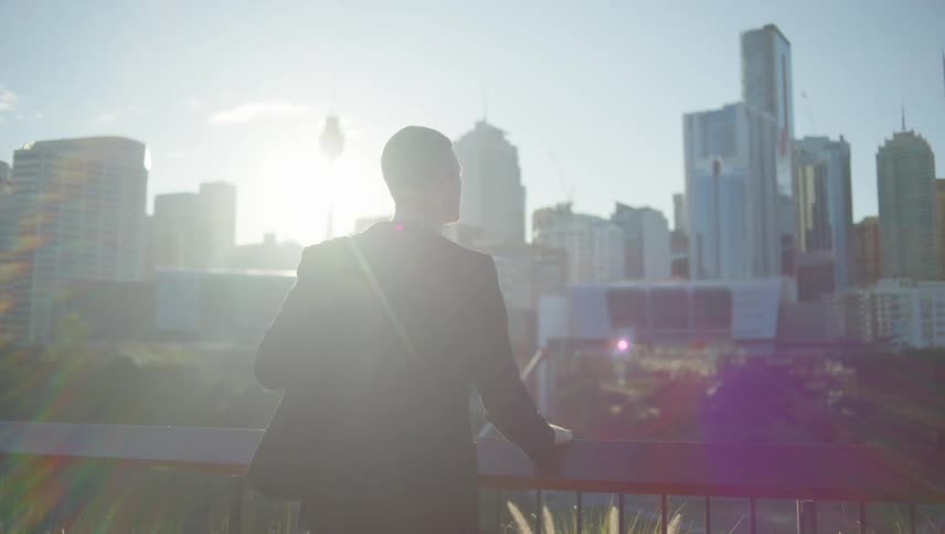 A man in formal wear stands on a balcony overlooking the city