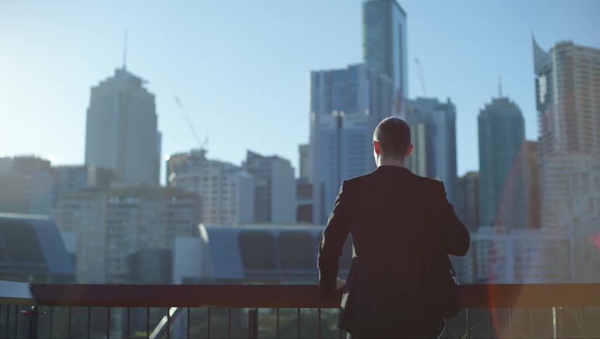 Man wearing a business suit stands at a rail looking at the city skyline on a sunny day