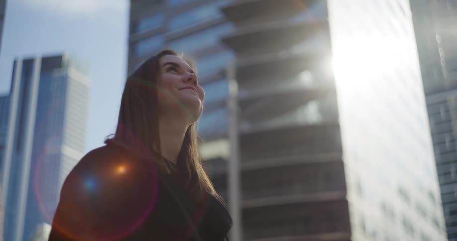 A happy womans face looking up and there are some big buildings around her and strong sunlight coming from above