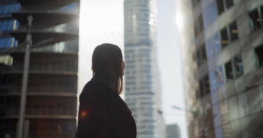 A woman is standing with her back turned and looking at some huge buildings in front of her