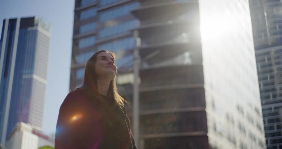 A woman looks up at the tall skyscrapers in the city on a sunny day