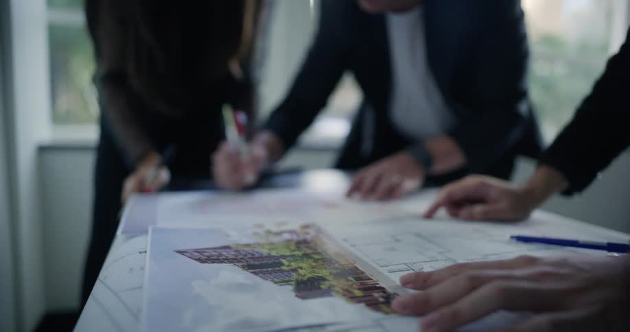 Three office employees write and point fingers over blueprints spread out on a table