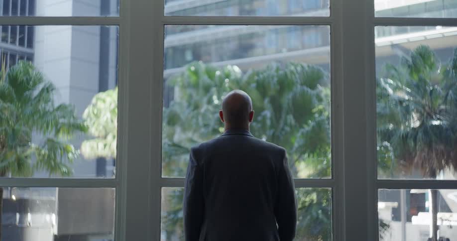 Businessman stands looking out the window of a downtown office building during a sunny day