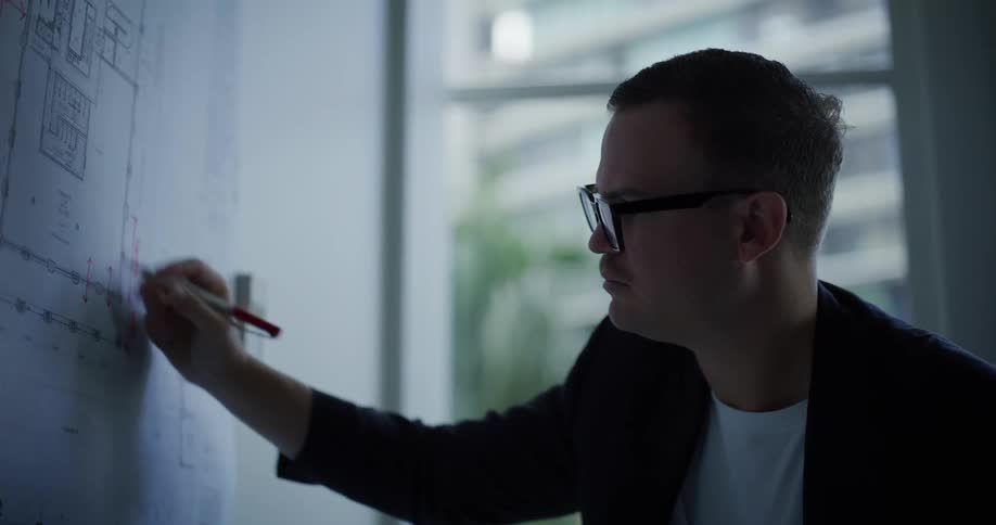 A man wearing glasses uses a pen to draw a sketch of a project on a piece of paper on the wall
