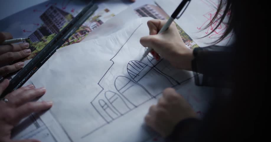 A woman using a pen to draw an arched building while two other people keep their hands on the table near the paper