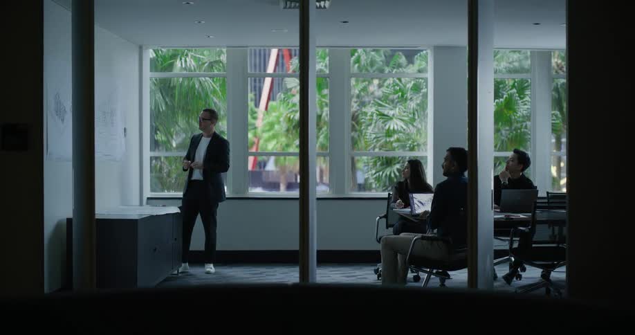 Man standing with a pen presenting a project to three people watching sitting at a table
