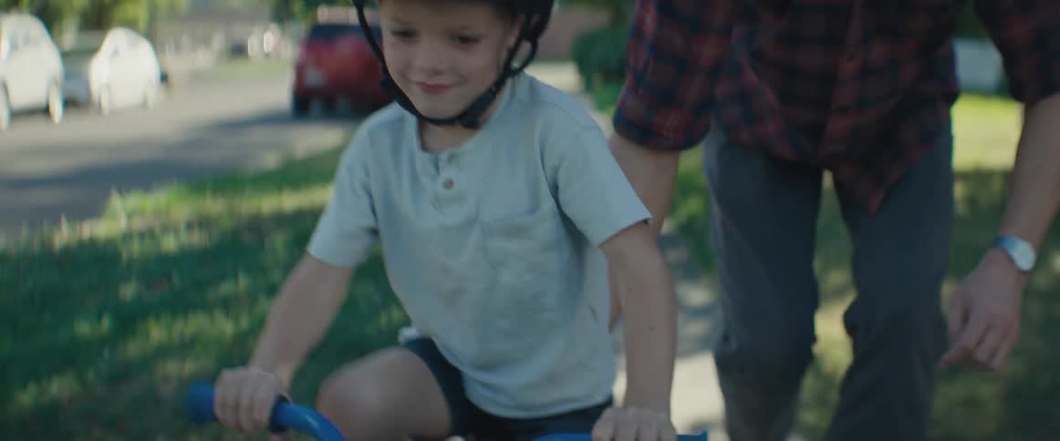 A young boy wearing a helmet learning to ride a bike with his father closely supporting him