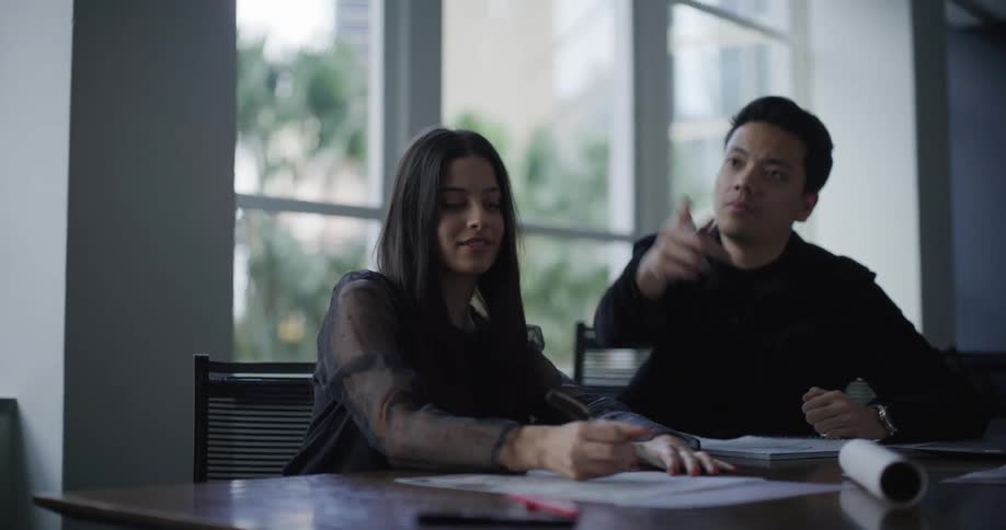 A woman and a man are seated at a table in a modern welllit room with the woman writing on a piece of paper while the man observes attentively