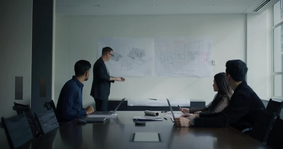 An office meeting in a large room with three people with laptops around a table while a fourth talks and writes on blueprints hanging on a wall