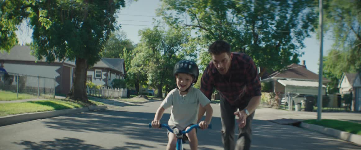 A young boy wearing a helmet learning to ride a bike with his father closely supporting him