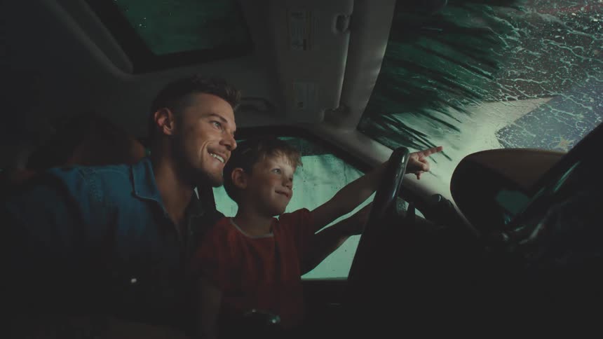 Father and son sitting in the same car seat watching the vehicle being washed by a machine
