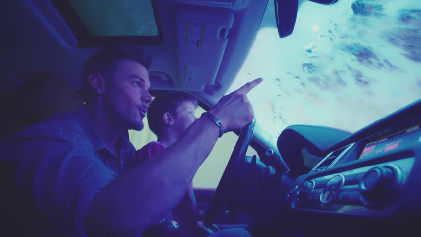 The son is sitting on his fathers lap while they drive in the rain