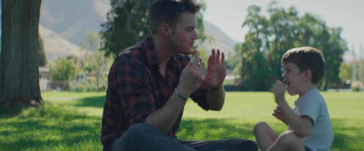 An adult man is sitting down and eating something while playing with his young son