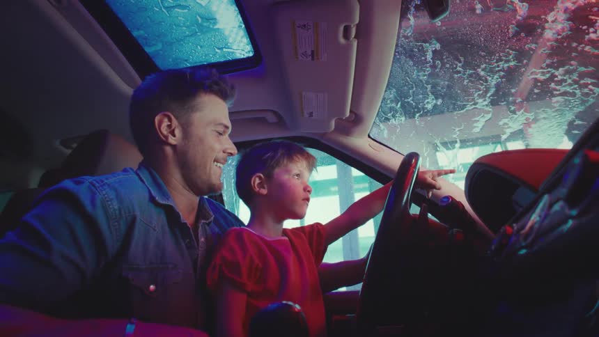 A man and his son are happily sitting inside a car while it is being washed on the outside