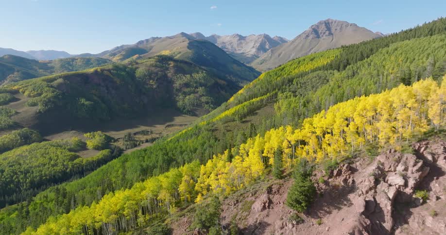 An aerial view from a drone captures a stunning mountain valley featuring a vibrant mix of green and golden trees with towering peaks and clear blue skies in the background