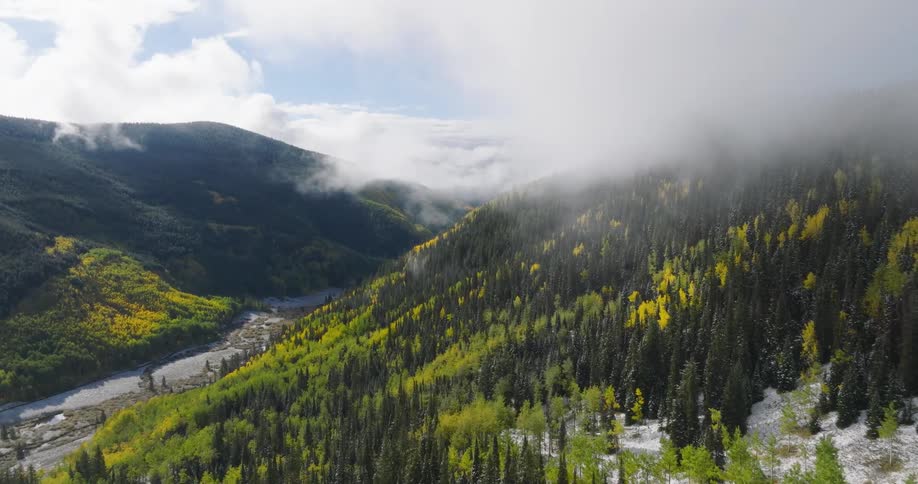 Aerial view of a mountainous landscape with a river winding through a valley surrounded by lush green forests dotted with patches of vibrant autumn foliage