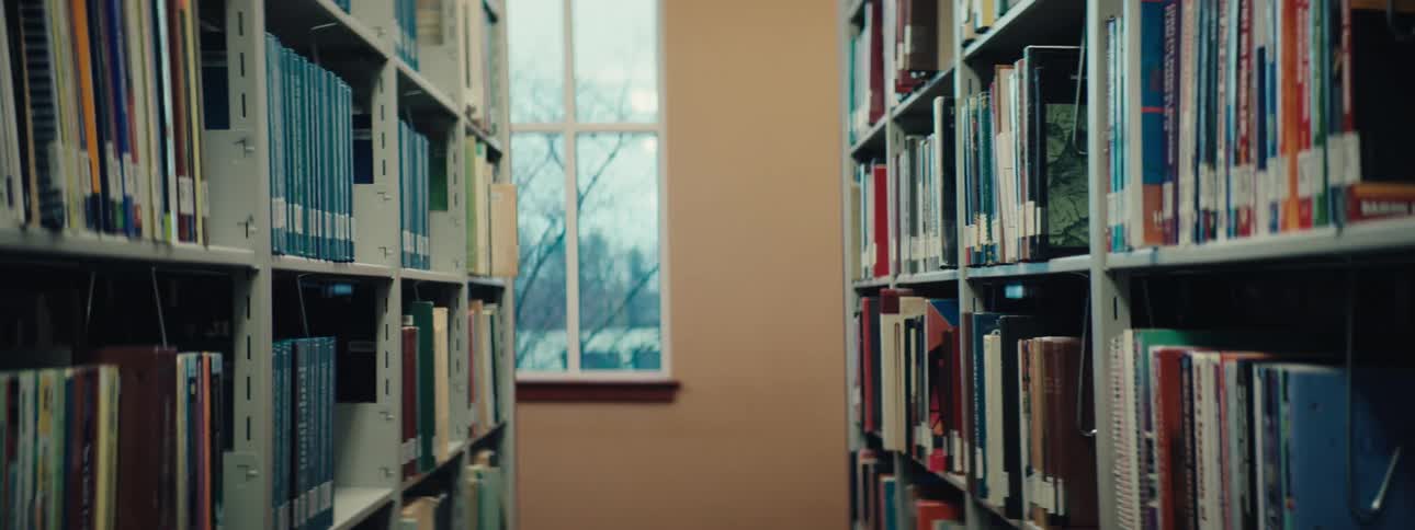 A library aisle with bookshelves on both sides and a window at the end