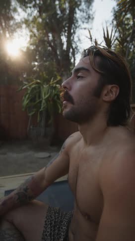 Man sitting in his backyard wipes his eyes and combs back his hair before looking around on a sunny summer day