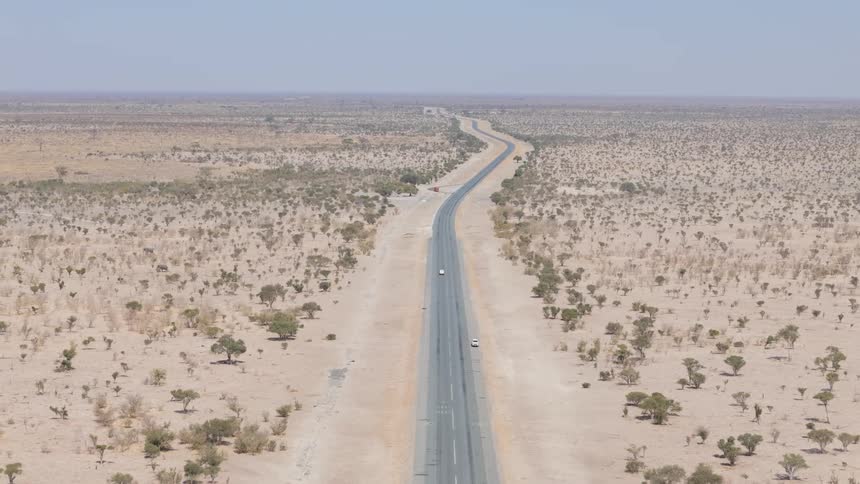 A white car drives on a straight empty road cutting through a vast dry landscape