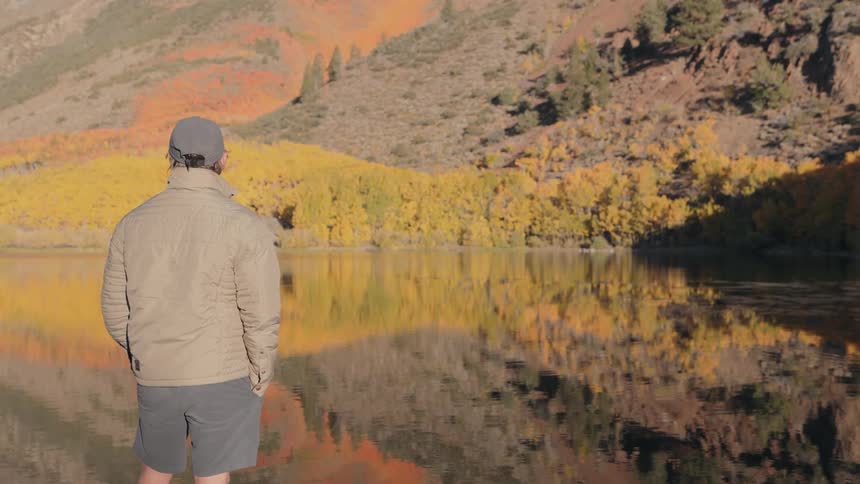 A man in a tan jacket and shorts stands on the edge of a lake gazing at the vibrant autumn foliage reflected on the water