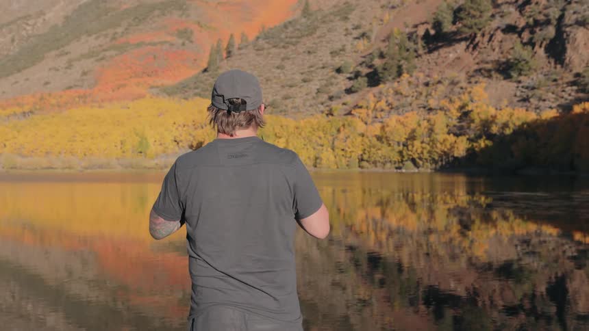 A white male puts on sunglasses while standing at the edge of a lake near a mountain