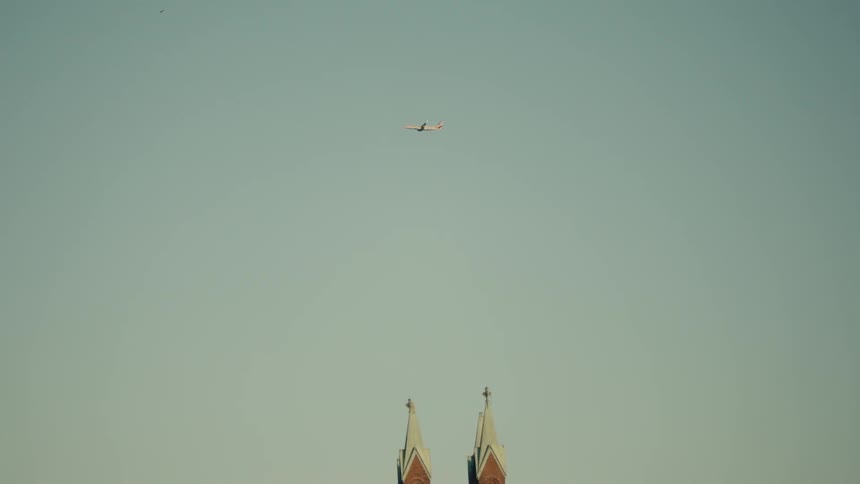 A plane flies high in the daytime sky above two spires on an unseen building