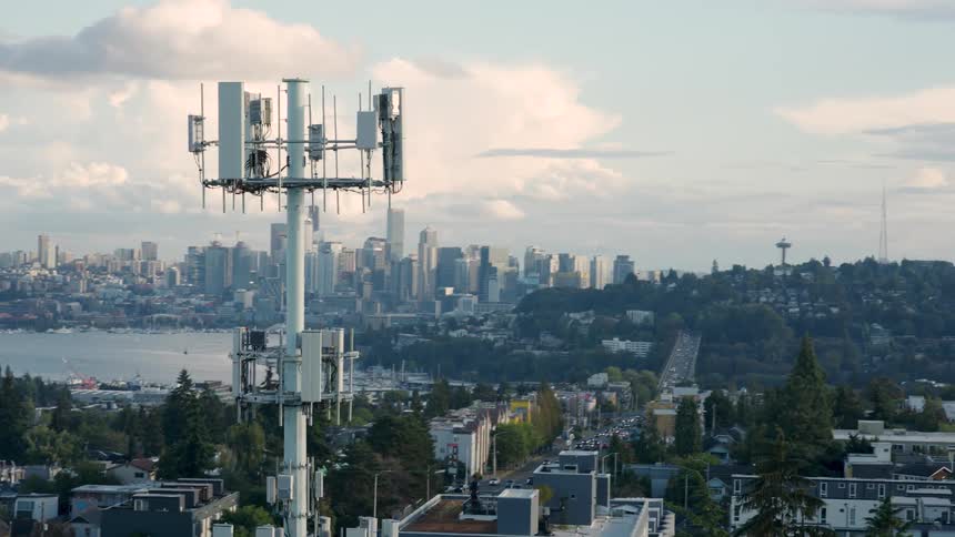 Descending orbit around a tall 5G communication tower atop a hill overlooking a sprawling urban cityscape