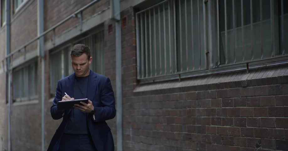 A man in a black suit stands on an alleyway drawing on a tablet or clipboard