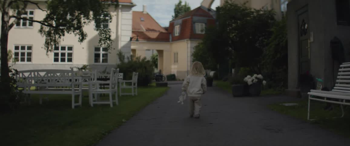 A little blonde girl in white clothes is walking on a path in a village yard